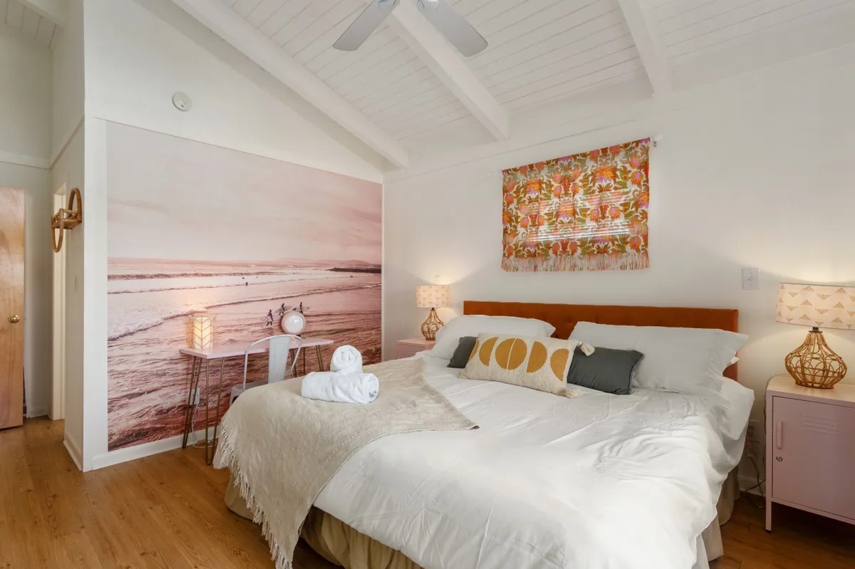 A spacious bedroom featuring a large bed with decorative pillows, an ocean-themed wall mural, a nightstand with a lamp, and colorful window treatments. The room is filled with natural light and exudes a calm, serene atmosphere, inviting relaxation after a day at the beach.