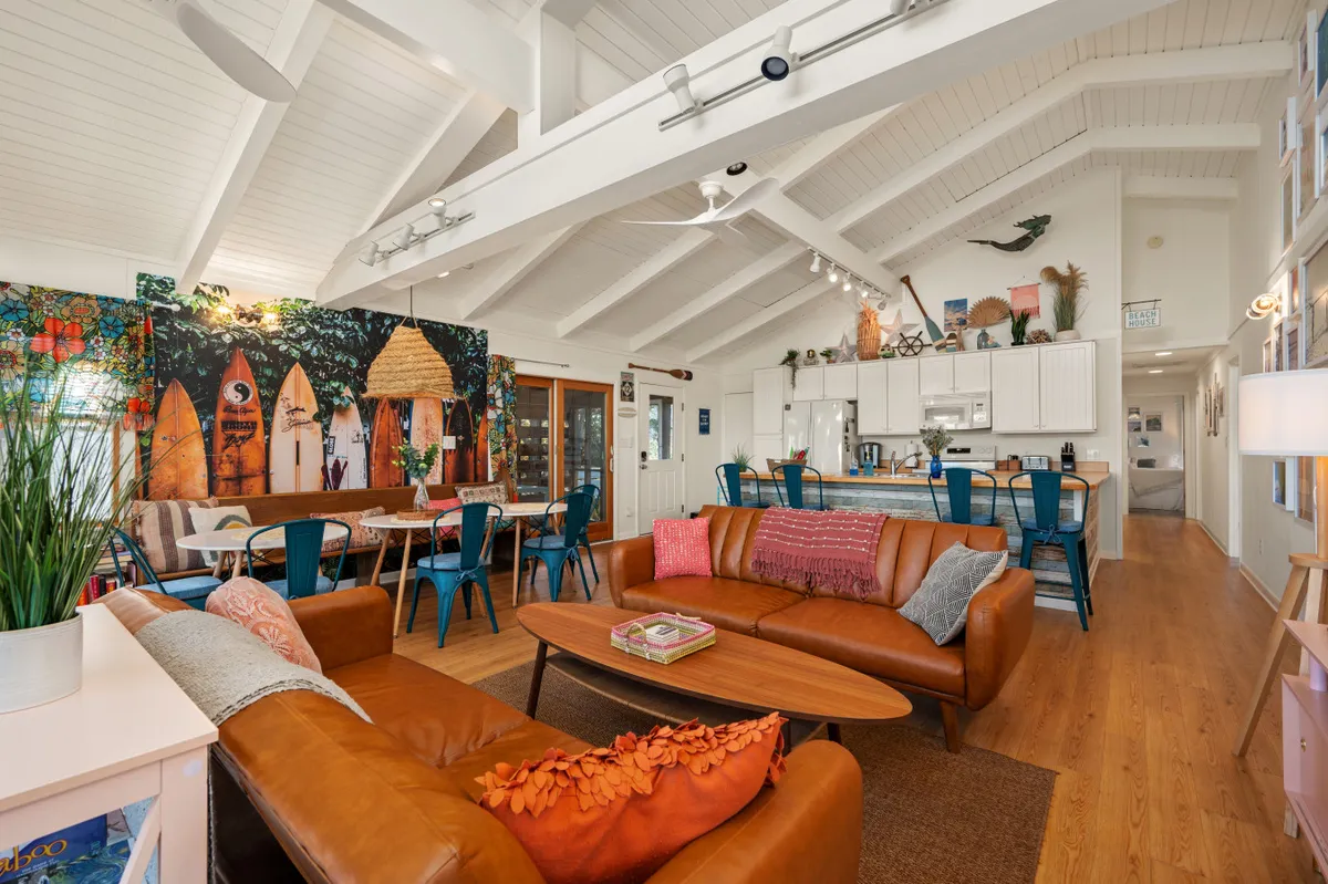 This image showcases a lively living and dining space with a colorful surfboard mural, a large wooden dining table surrounded by bright blue chairs, and a comfortable leather sofa. The area is well-lit by natural light streaming through expansive windows, highlighting the open floor plan and coastal-inspired décor.