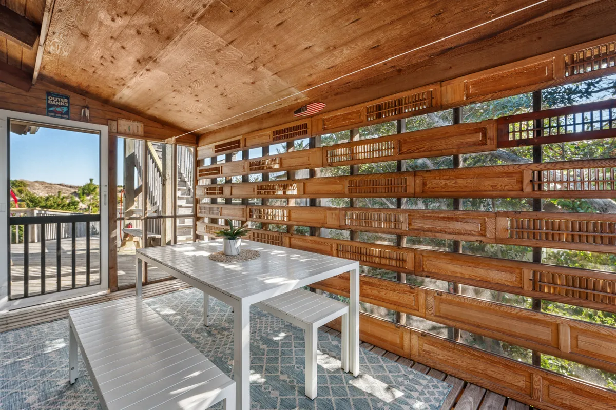 A cozy screened porch with wooden slatted walls, a white table and benches, and a decorative plant centerpiece. The space is airy and inviting, offering a stylish spot for outdoor dining and relaxation with views of greenery.