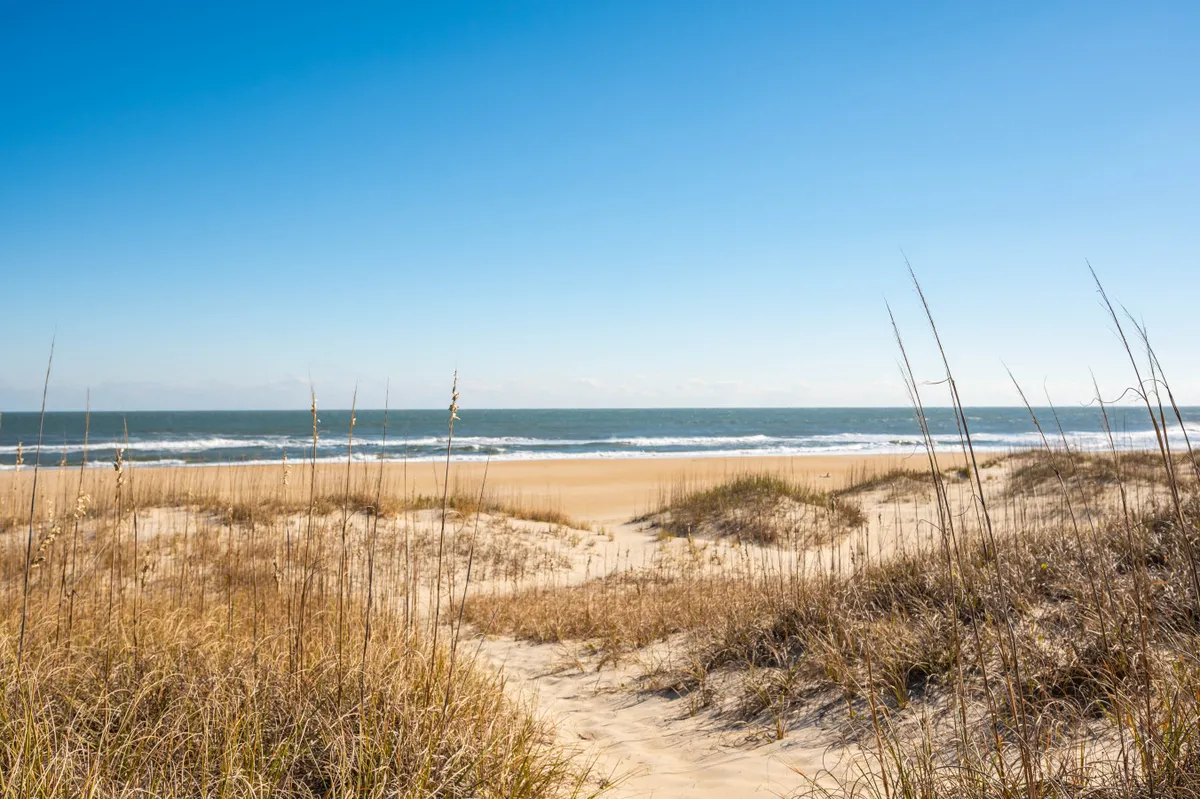 A serene sandy beach with rolling waves and dune grass, bathed in natural light. The beach appears inviting, perfect for walks, sunbathing, or catching sunrise over the Atlantic.