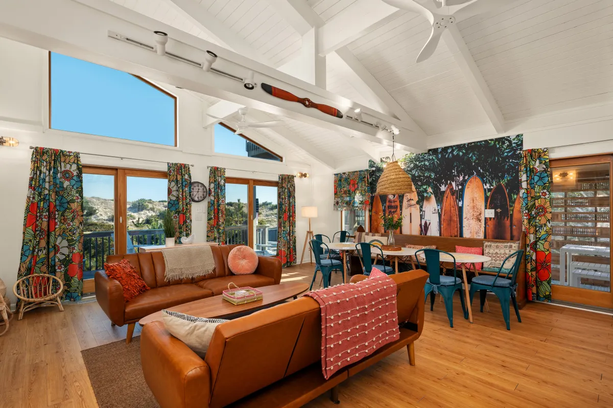 A spacious interior featuring large windows with sunset views, colorful curtains, a wooden dining table, a vibrant surfboard mural, and a leather sofa. The space is flooded with natural light and offers a seamless transition from the living area to the deck beyond.