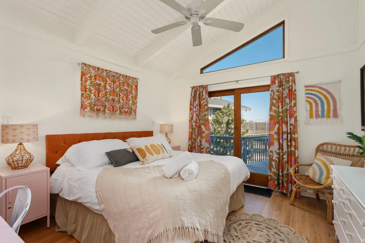 A cozy bedroom with a bed dressed in colorful bedding, a large window letting in sunshine, decorative curtains, and a nightstand with a lamp. The intimate setting is inviting for restful nights or peaceful mornings.