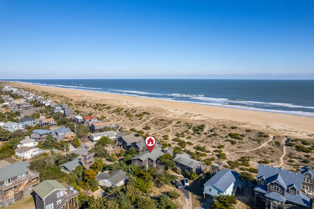 The image showcases the exterior of a single-family beach house set against the sweeping dunes of Avon, North Carolina. The foreground features a path leading through dune grass directly to an expansive, uncrowded beachfront with the Atlantic Ocean shimmering in the background. Neighboring beach homes dot the tranquil landscape, while the early morning or late afternoon light suggests a serene, inviting atmosphere perfect for leisurely walks, beach bonfires, and ocean adventures.
