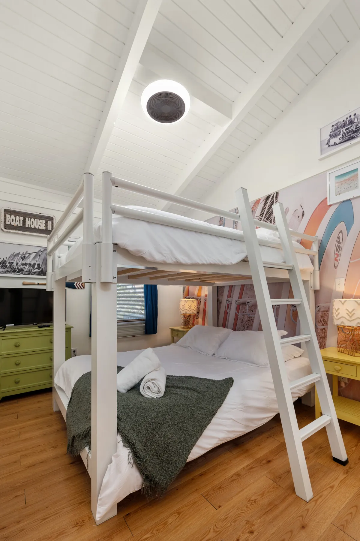 A cozy bunk room with a queen bunk bed, vibrant wall art, a green dresser, and warm lamp lighting. The playful yet comfortable setting is ideal for kids or teens, offering a fun yet restful space for sleep and creativity.