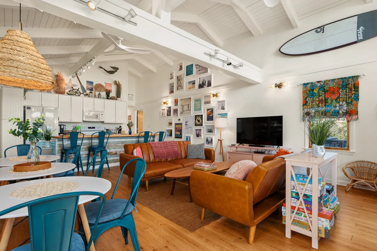 Featuring a cozy sofa seating area, a kitchen with bar stools, a gallery wall of photographs and art, colorful window treatments, and a surfboard on the wall. The space is bright and inviting, ideal for family gatherings, game nights, and casual dining.
