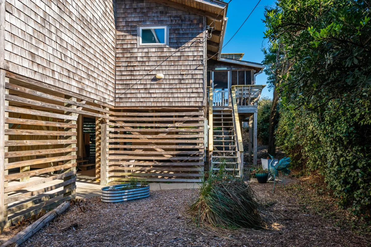 This image captures the exterior of the property, featuring wooden siding, stairs leading to an upper deck, textured ground cover, and small plants. The photo conveys a welcoming outdoor gathering area that is ideal for hosting barbecues or relaxing with friends.