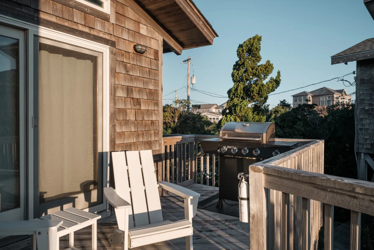 An exterior deck area featuring a BBQ grill, Adirondack chair, and views of surrounding houses. The area is set up for outdoor cooking and relaxation, inviting guests to savor grilled meals in the fresh coastal air.