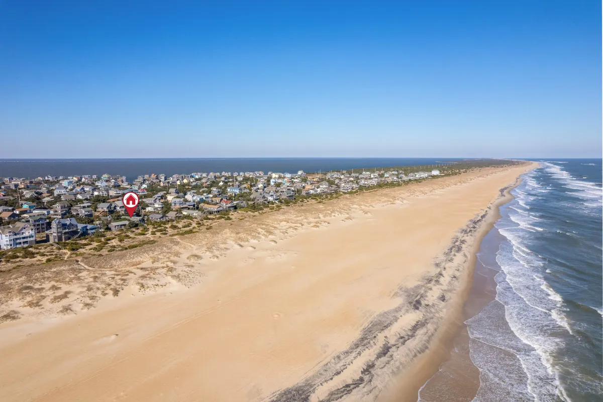 A wide view of the beach with gentle ocean waves, sand dunes in the foreground, and several houses nestled along the coastline. The scene is lively yet tranquil, hinting at water-based activities such as surfing and beach gatherings.