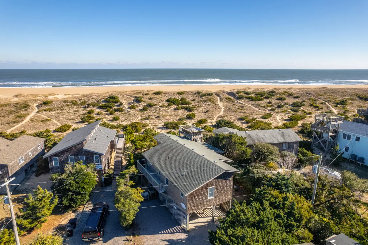 A wide beachfront scene with rolling sand dunes, ocean views, and the silhouette of nearby houses. The setting is tranquil, with hints of recent activity like footprints in the sand, suggesting family walks and gatherings at the beach.