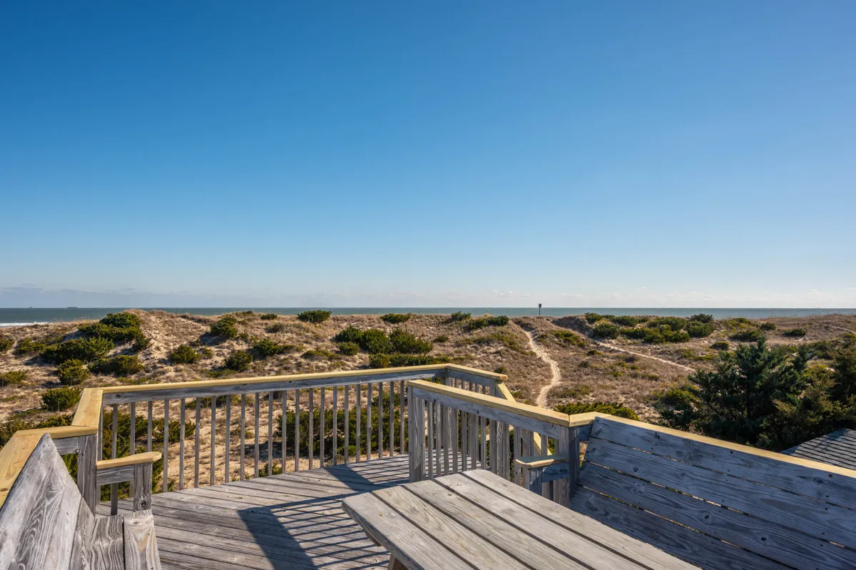 A serene view from a wooden deck overlooking the dune landscape and ocean beyond. The deck is an inviting spot for relaxing with morning coffee, watching sunsets, or unwinding after a day at the beach.